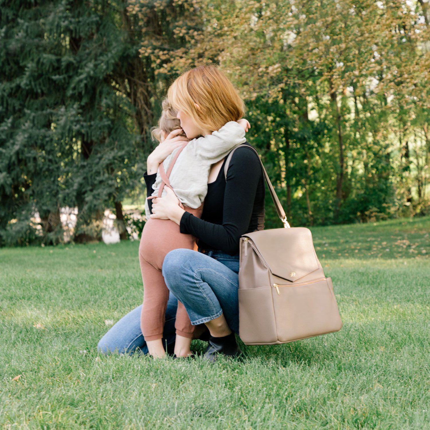 A parent hugs their child while wearing the Aspen Diaper Bag II in stone.