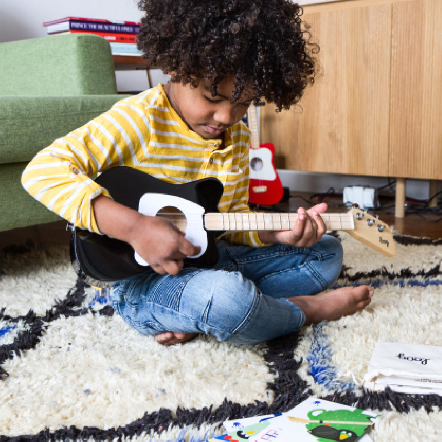 Child playing a black Loog Mini acoustic guitar on a rug