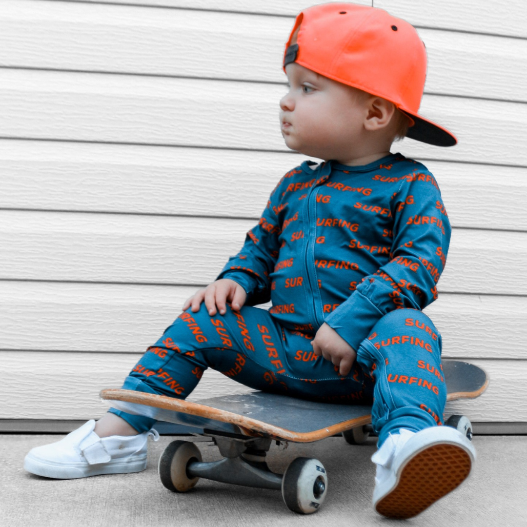 Toddler in Pipeline Dreams surfing print sleeper sitting on a skateboard.