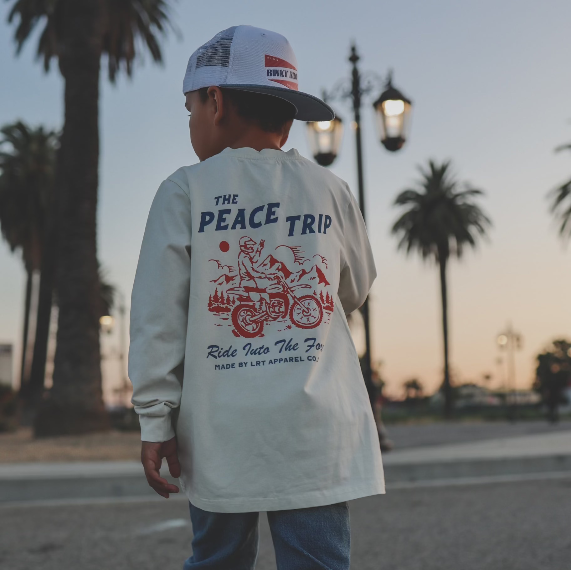 Boy wearing 'Peace Trip' long sleeve tee, back view, palm tree background.