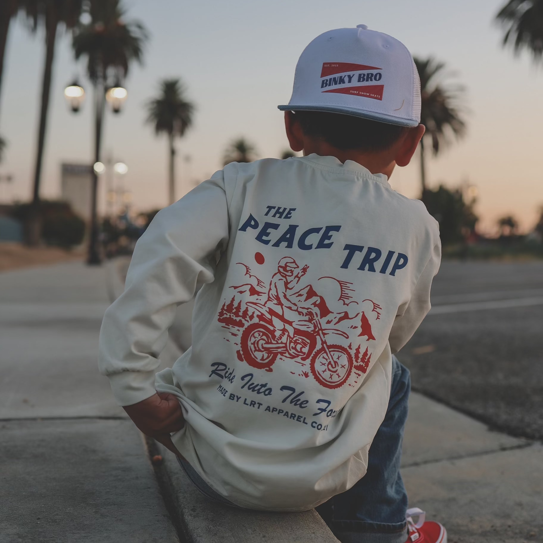 Child in 'Peace Trip' white long sleeve tee, red checkered sleeves, palm trees.