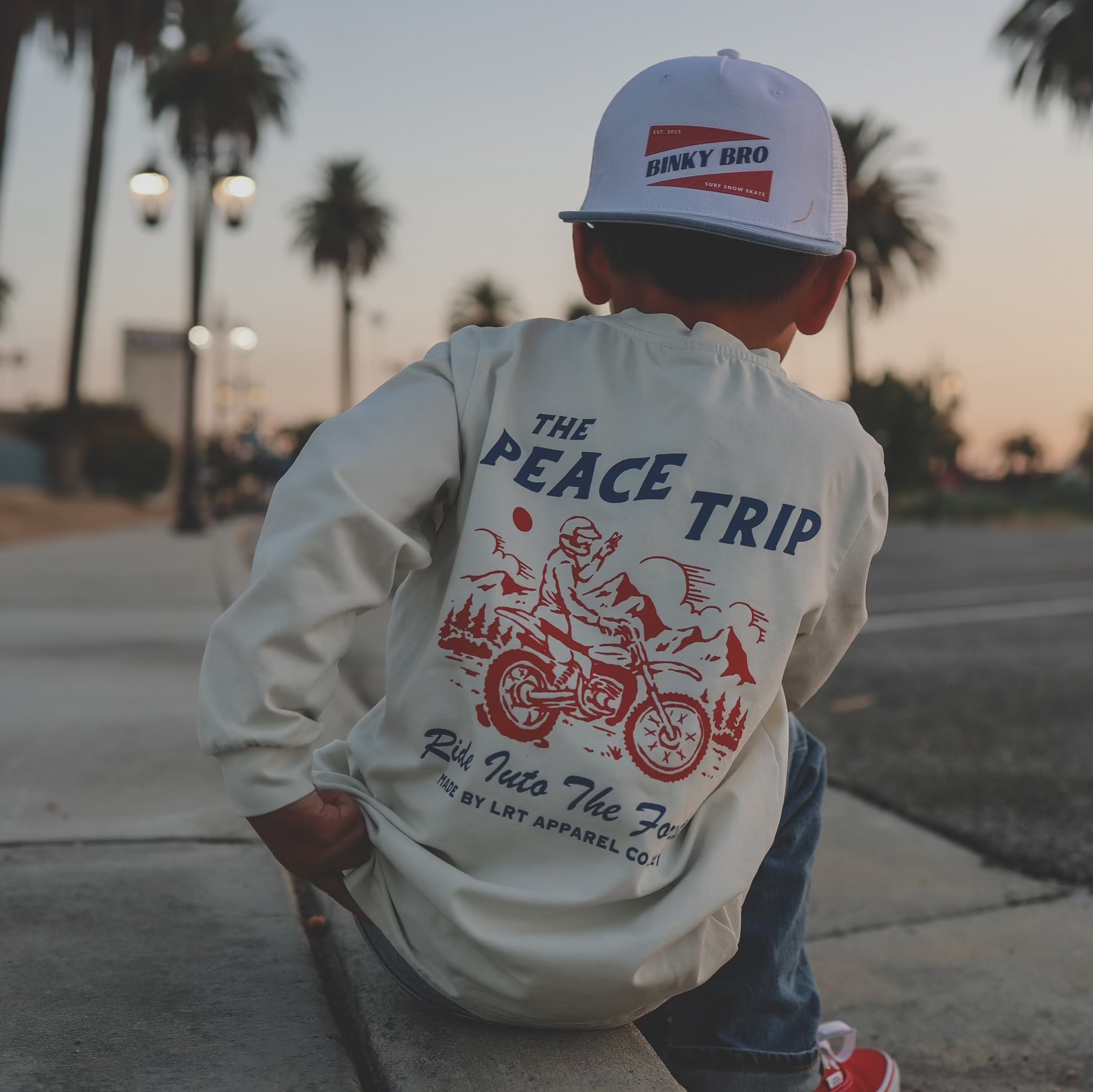 Child in 'Peace Trip' white long sleeve tee, red checkered sleeves, palm trees.