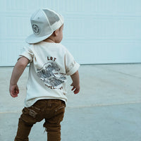 Child in Swell Days Tee, brown pants and hat, playing outside.