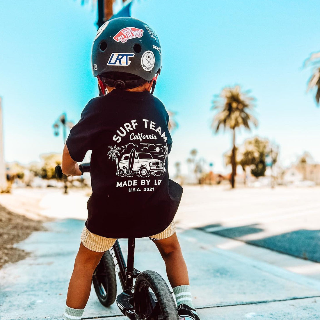 Child on bike wearing Surf Team tee and helmet, back view.