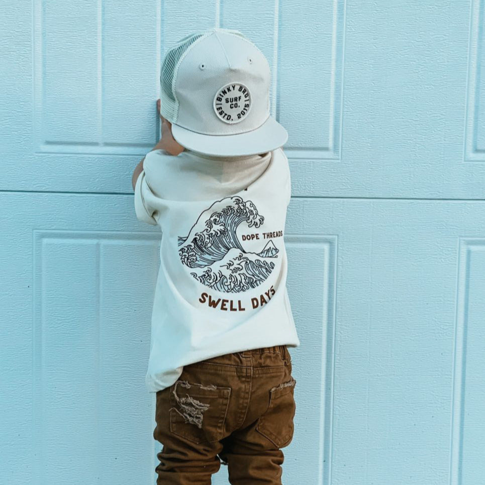 Toddler wearing cream Swell Days Tee and trucker hat, back view.