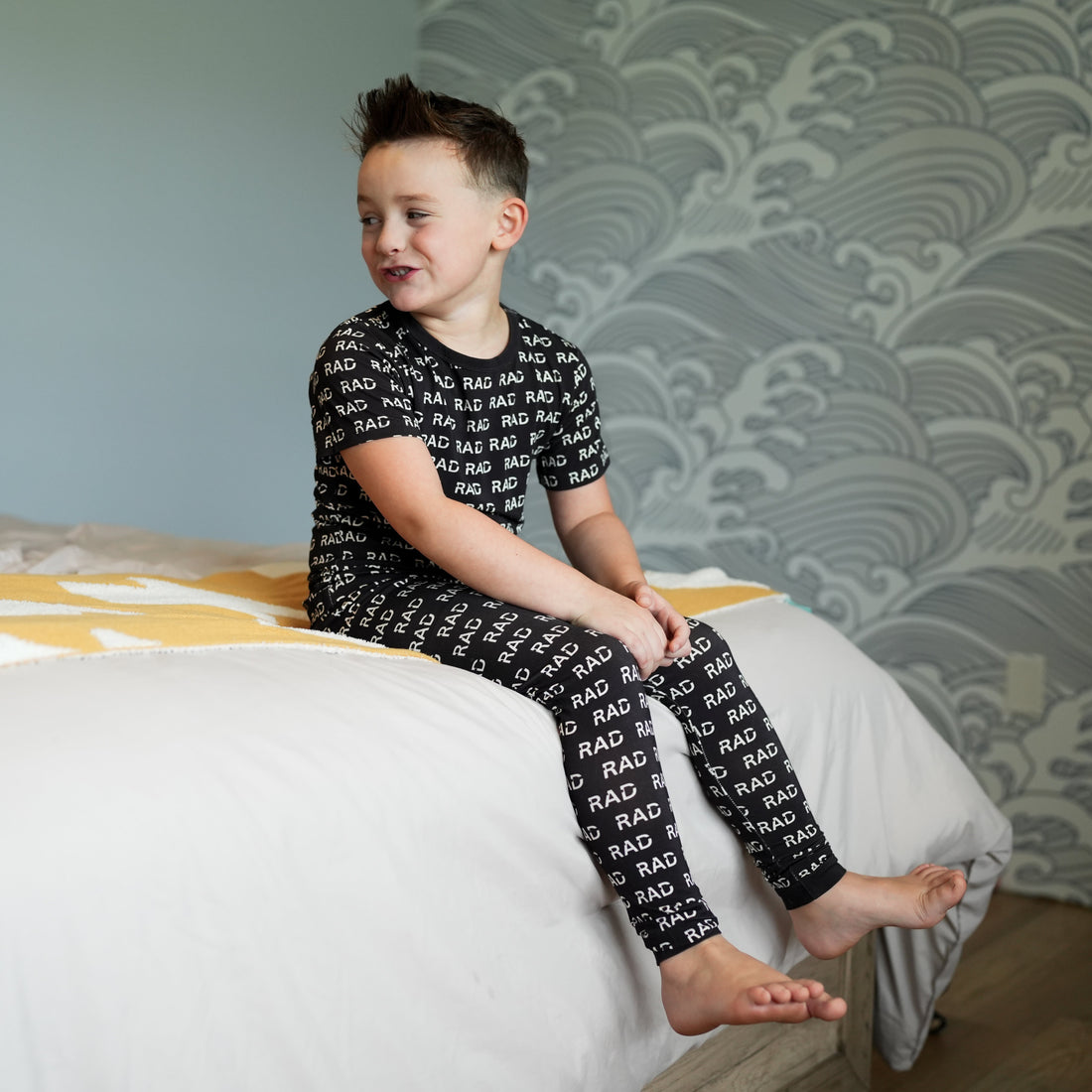 Boy in black RAD print bamboo pajamas sitting on bed.