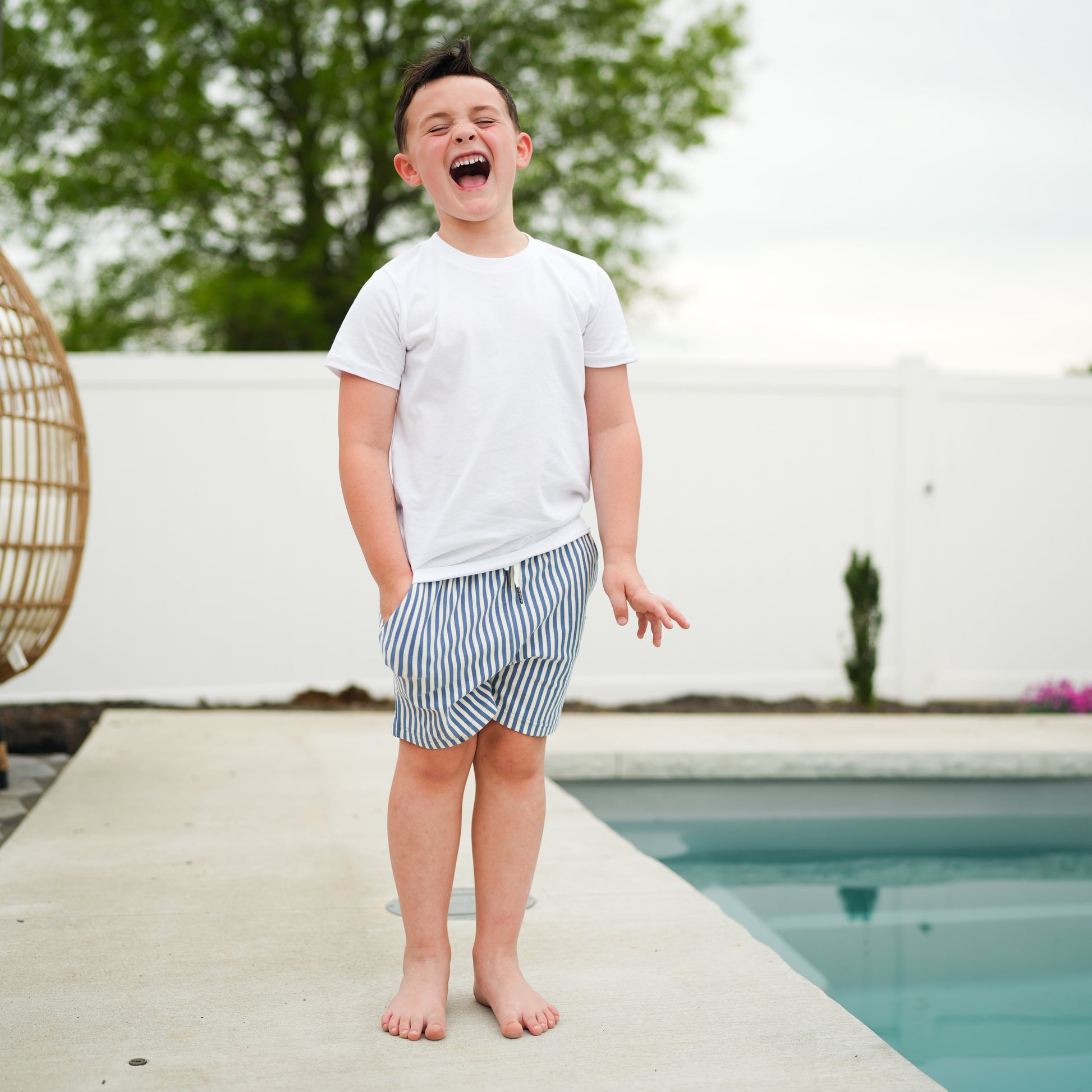 Boy laughing by pool in RAD Organics white tee and striped swim trunks.