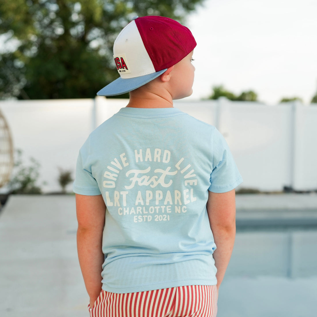 Boy wearing cloud blue APEX LIVE FAST tee, back view, with hat and shorts.