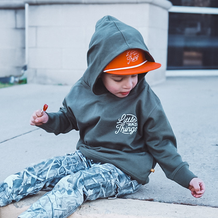 Child in army green Radiator Hoodie sitting and holding a lollipop.