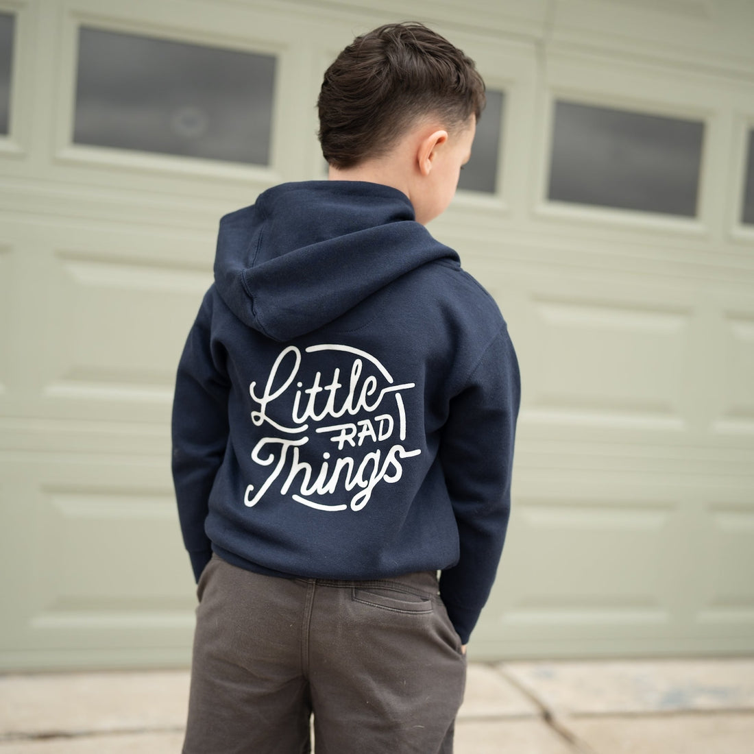 Child in navy Radiator Hoodie with 'Little Rad Things' design, standing in front of a garage.