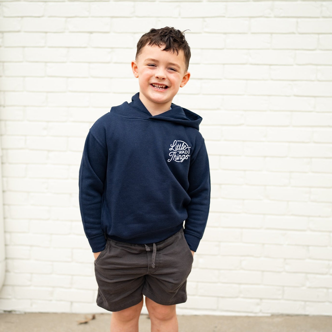 Smiling boy in navy Radiator Hoodie with logo, standing against a white brick wall.