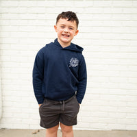 Smiling boy in navy Radiator Hoodie with logo, standing against a white brick wall.