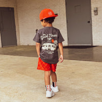 Child in red/orange checkerboard Retro-Wave hybrid board shorts holding a toy.