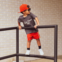 Child in red/orange checkerboard Retro-Wave hybrid board shorts leaning on a rail.