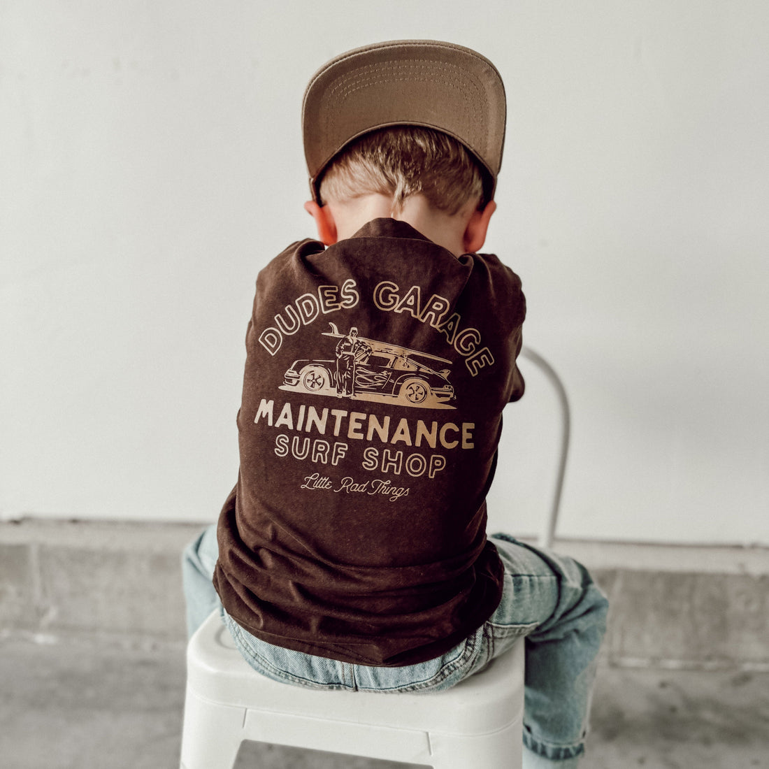 Child in Dudes Garage brown t-shirt with retro car graphic on a white stool.