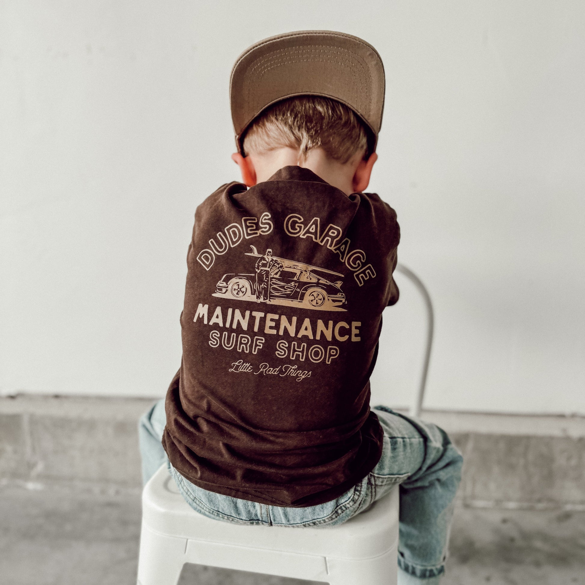 Child in Dudes Garage brown t-shirt with retro car graphic on a white stool.