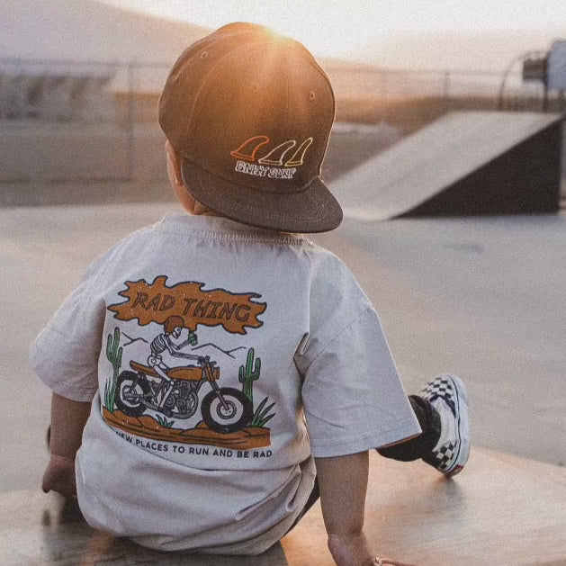 Toddler in 'Where the Rad Things Are' tee and cap, sitting at a skatepark.