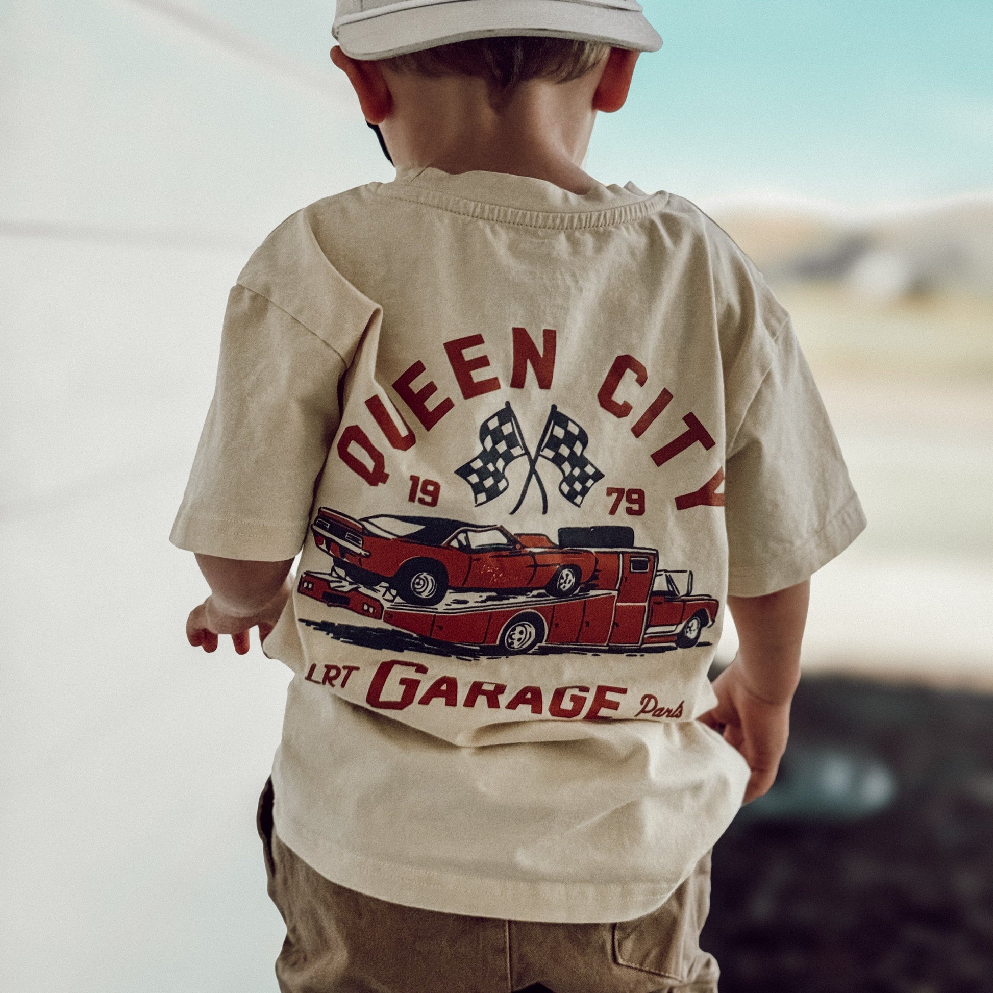 Toddler wearing Queen City Garage Tee, vintage cream with racing graphic.