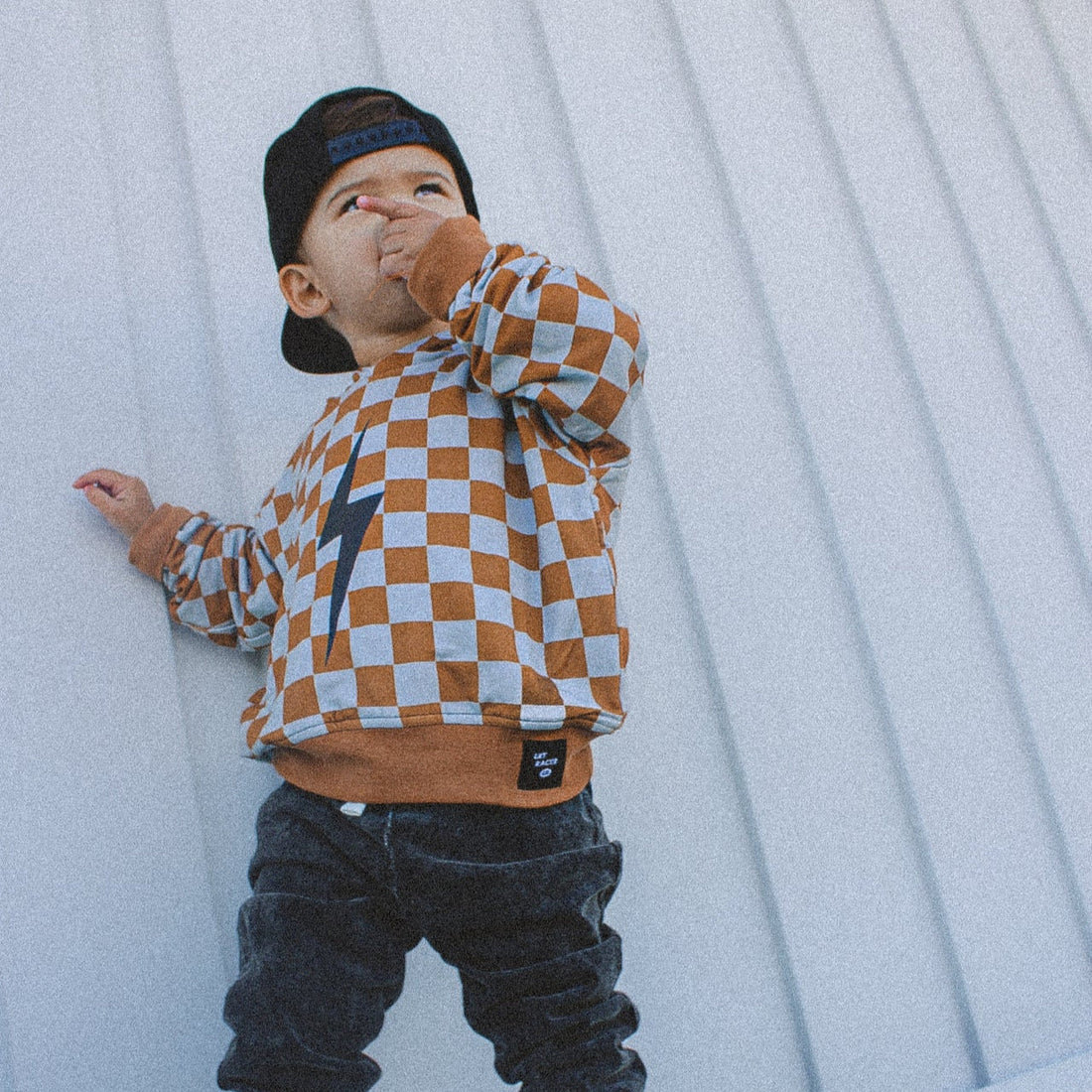 Little boy in a rust checkered hoodie and black jeans leaning against a wall.