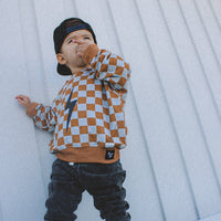 Little boy in a rust checkered hoodie and black jeans leaning against a wall.