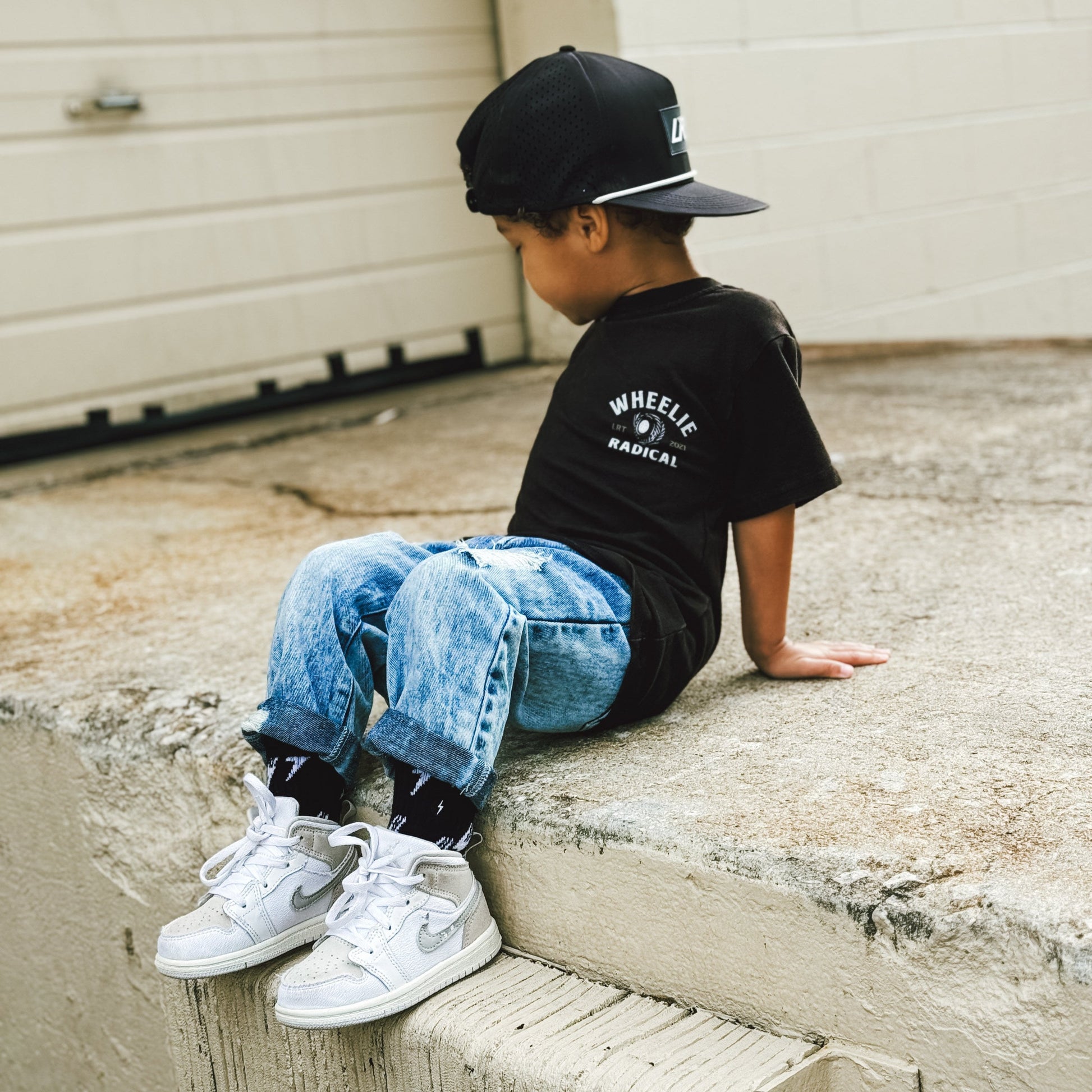 Child sitting wearing Wheelie Radical Tee with a monster truck and car graphic.