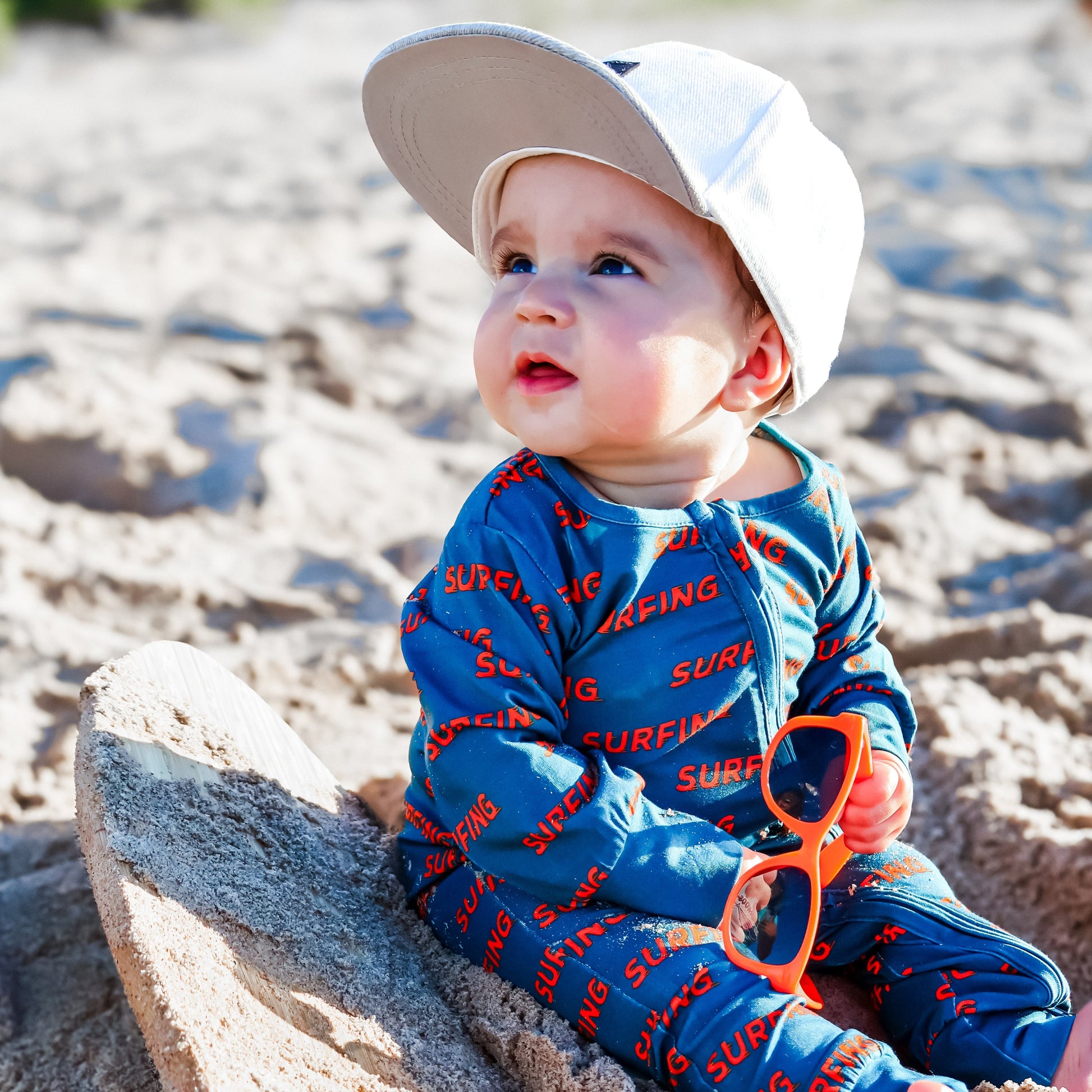 Baby in Pipeline Dreams surfer print sleeper with white hat and orange sunglasses.