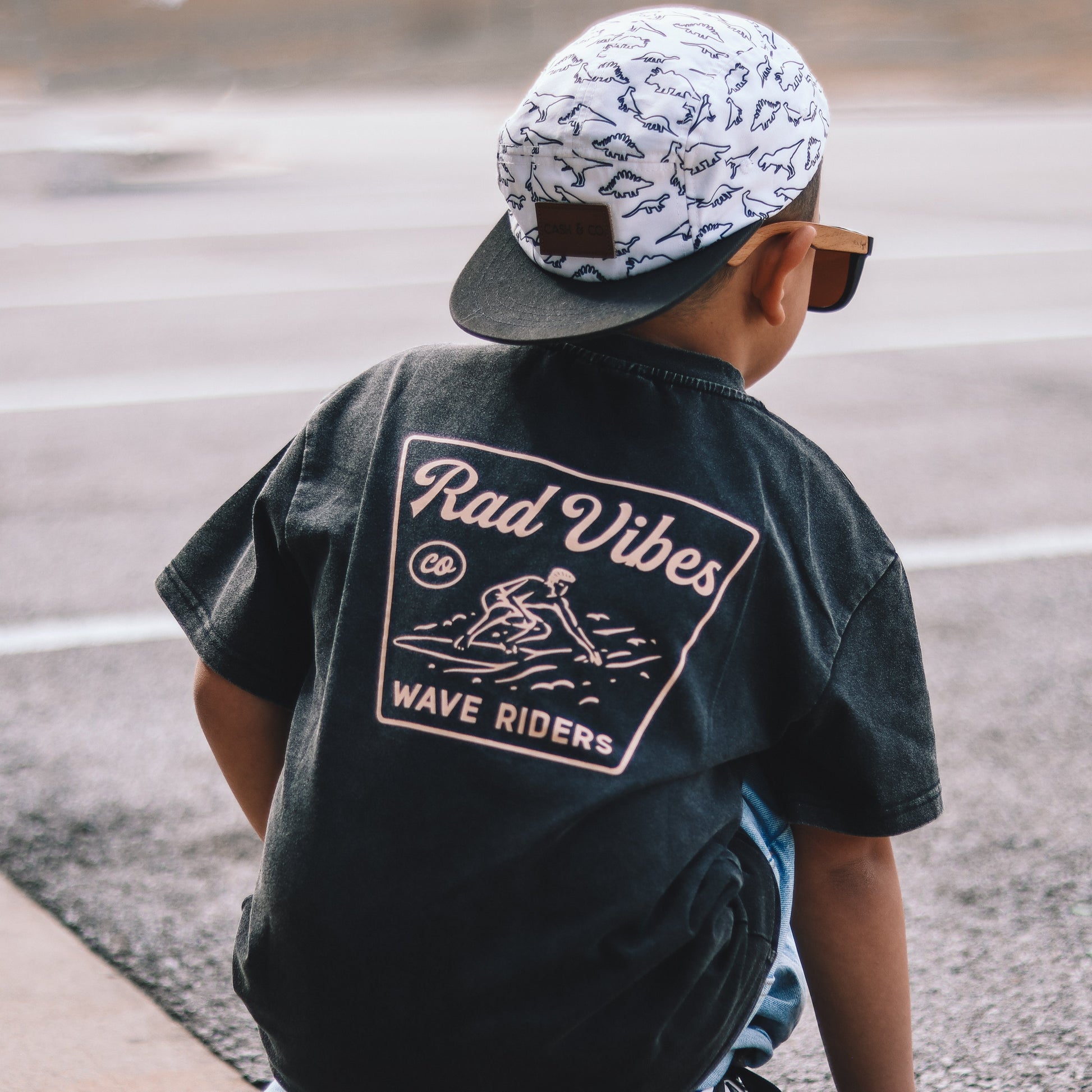 Child in Mavericks Tee vintage black and dinosaur hat sitting down.