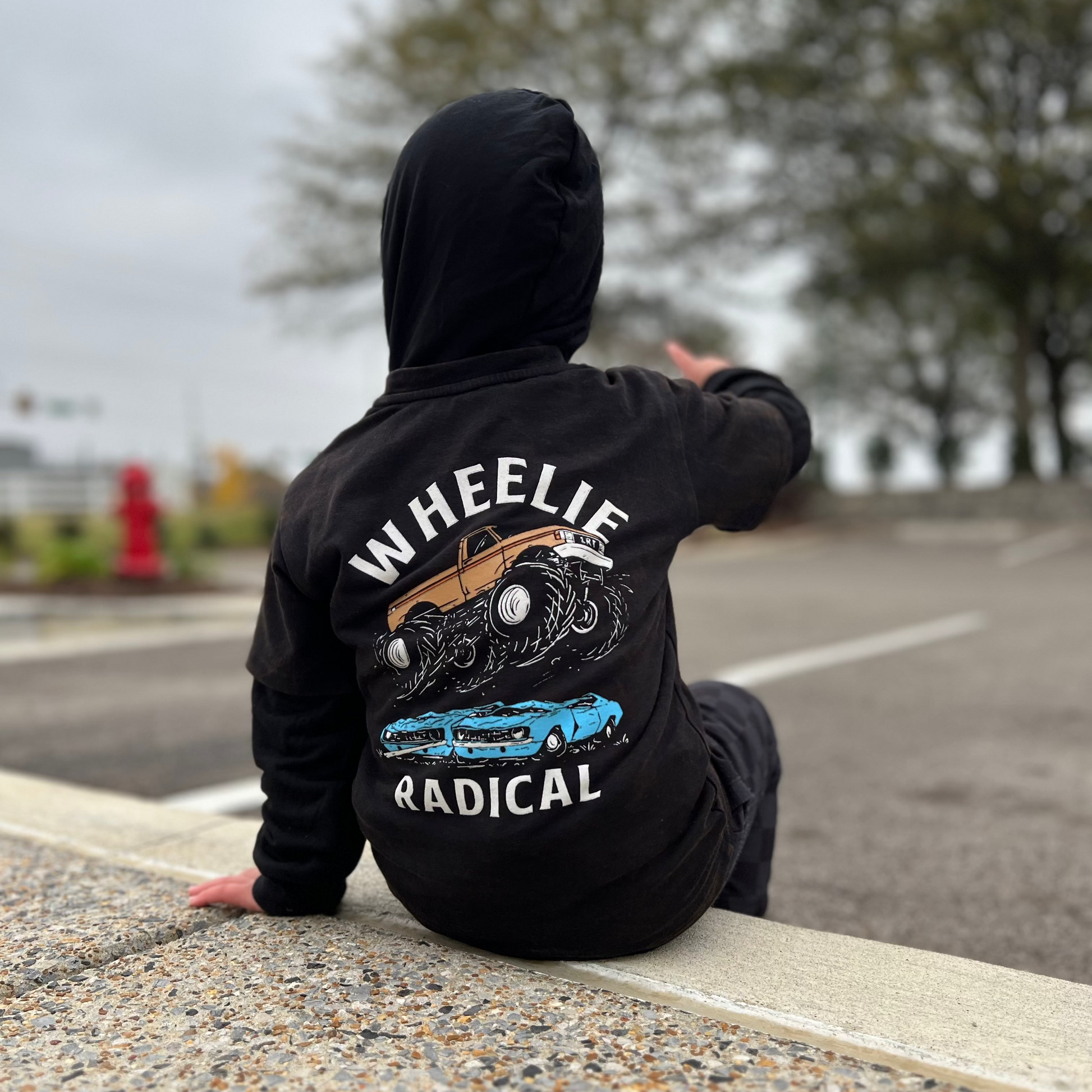 Boy sitting wearing Wheelie Radical Tee with a monster truck and car graphic.