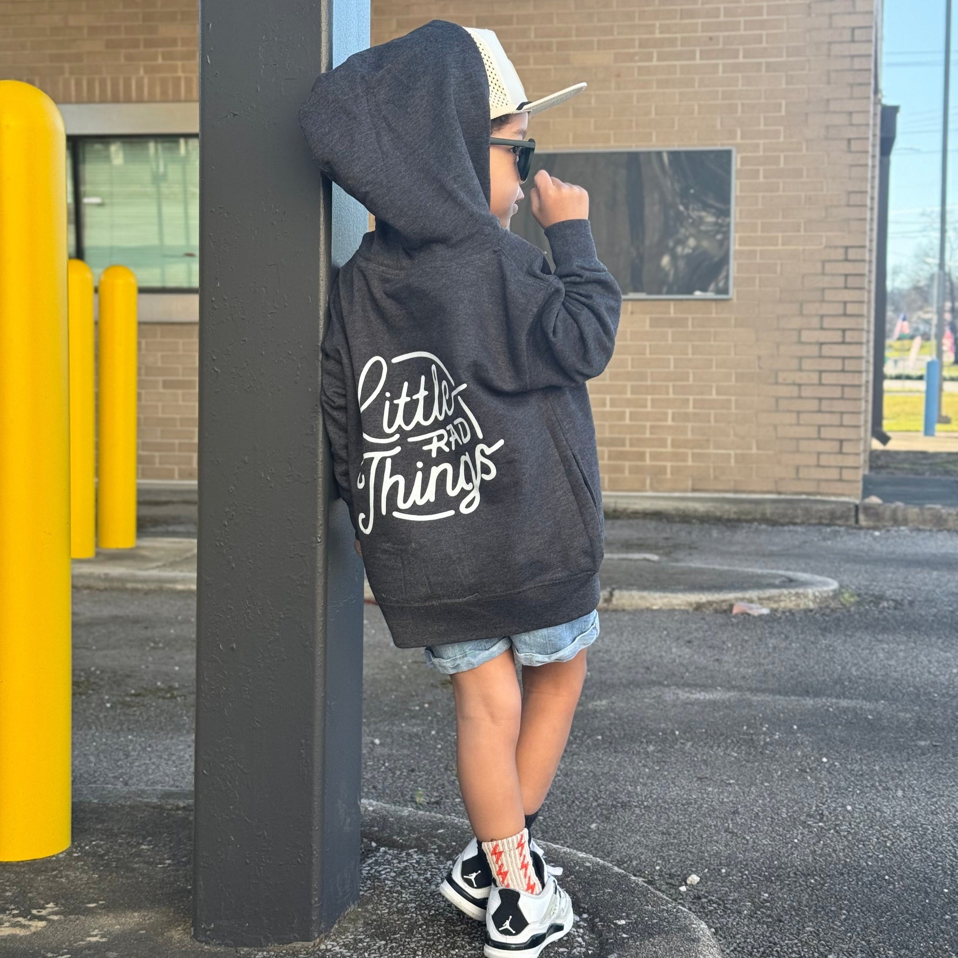 Smiling child in grey Radiator Hoodie, leaning against yellow poles.