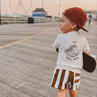 Little boy on boardwalk wearing Swell Days Tee and carrying skateboard.