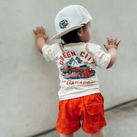 Toddler wearing orange checkerboard board shorts and a 'Queen City' t-shirt.