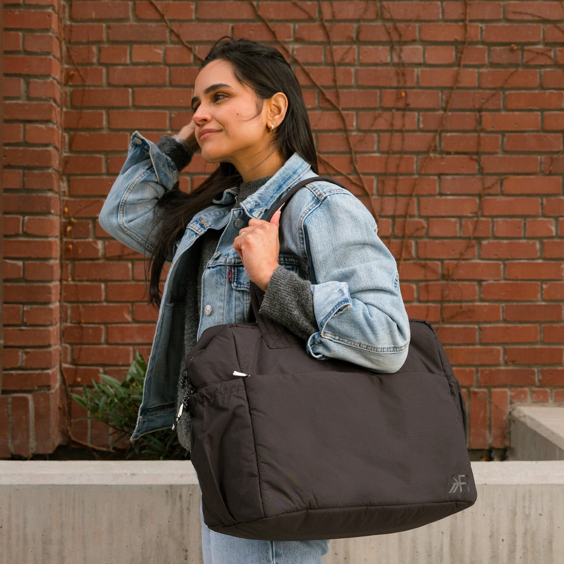 Woman carrying the Jet Motion Overnight Duffle in black with brick wall background.