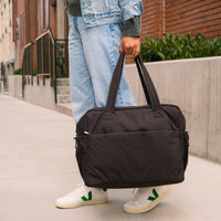 Jet Motion Overnight Duffle in black with top handles and shoulder strap, 'NEW!' badge.