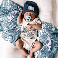 Baby in cream Dope Threads Swell Days organic cotton onesie with wave graphic.