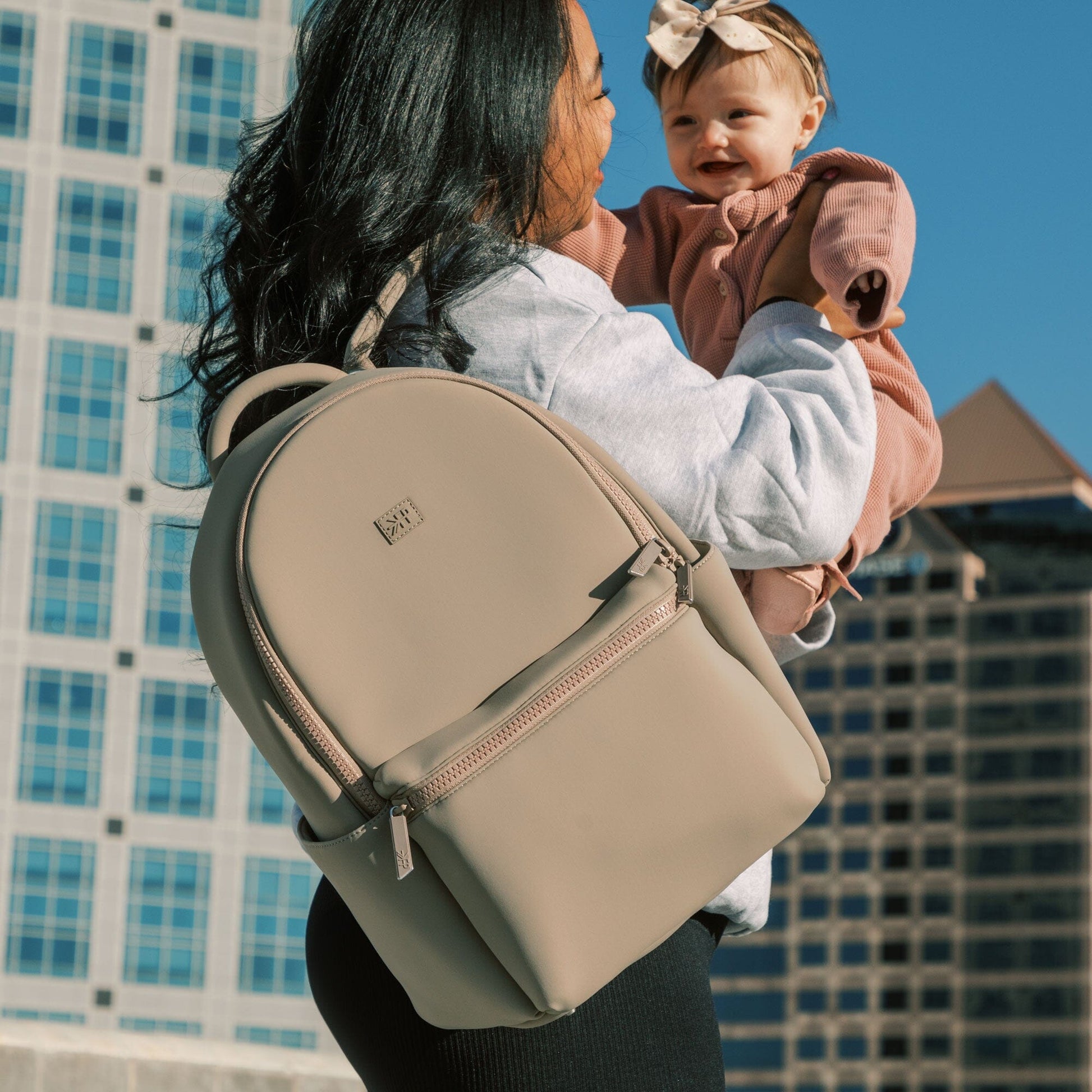 Mom holding baby with Aspen Seoul Neoprene Backpack in Sand.