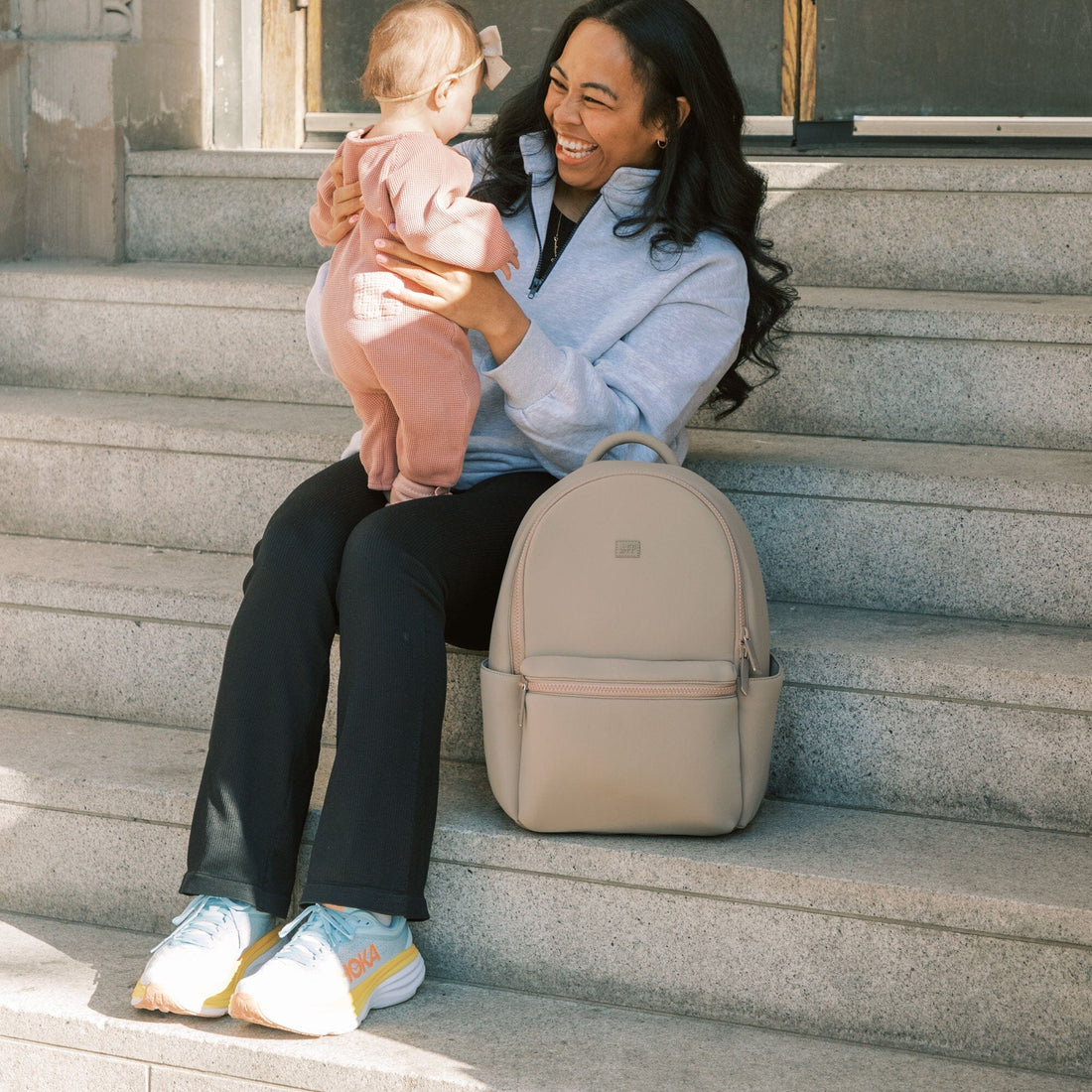 Mom with baby sitting next to the Aspen Seoul Backpack in Sand.