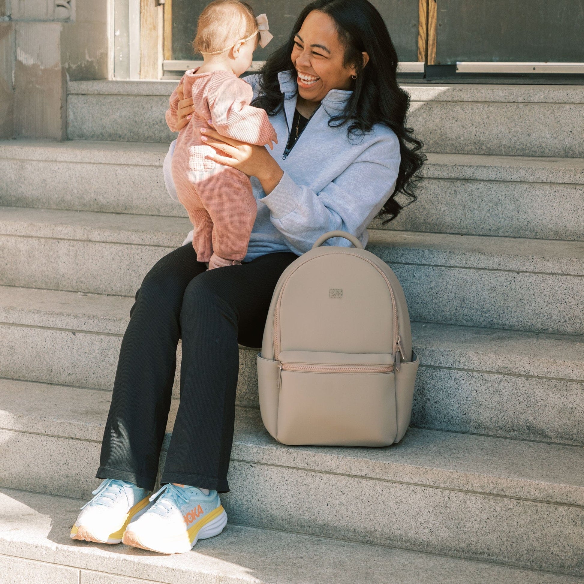 Mom with baby sitting next to the Aspen Seoul Backpack in Sand.