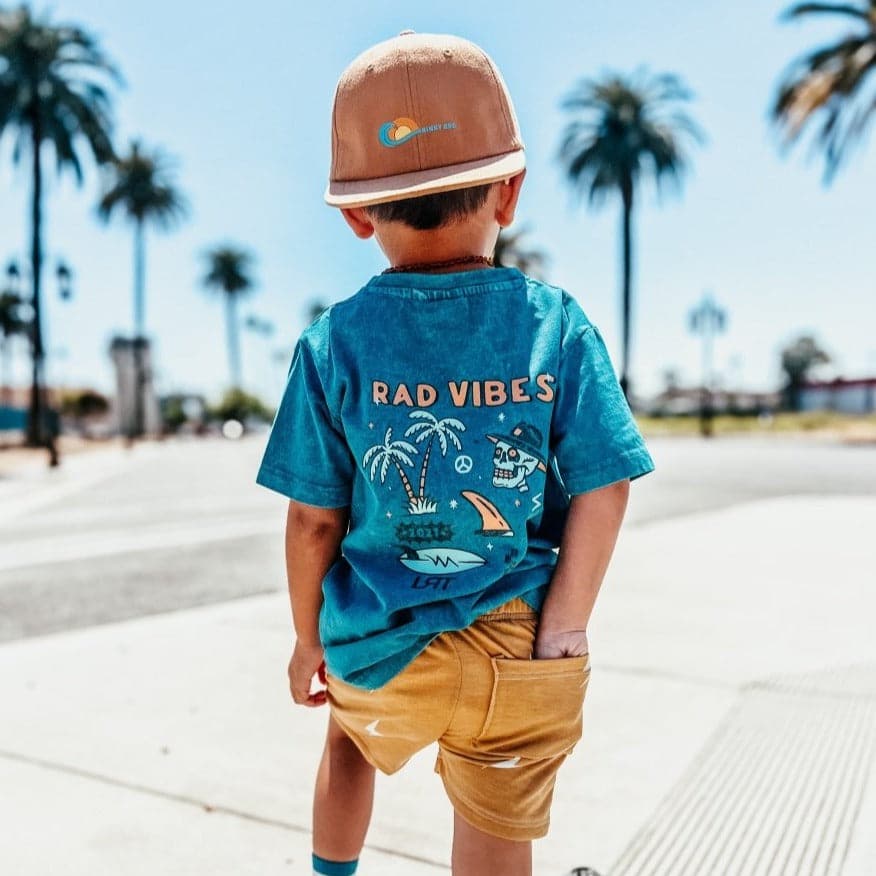 Boy wearing 'Rad Vibes' tee and gold shorts by palm trees