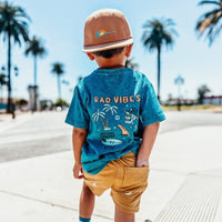 Boy wearing 'Rad Vibes' tee and gold shorts by palm trees