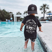 Young child in pool wearing Surf Team tee and matching swim trunks.